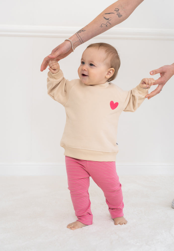 2-teiliges Baby Set mit beigem Sweatshirt mit pinkem Herz-Print und pink-rosa gestreifter Leggings, flach auf weißem Hintergrund