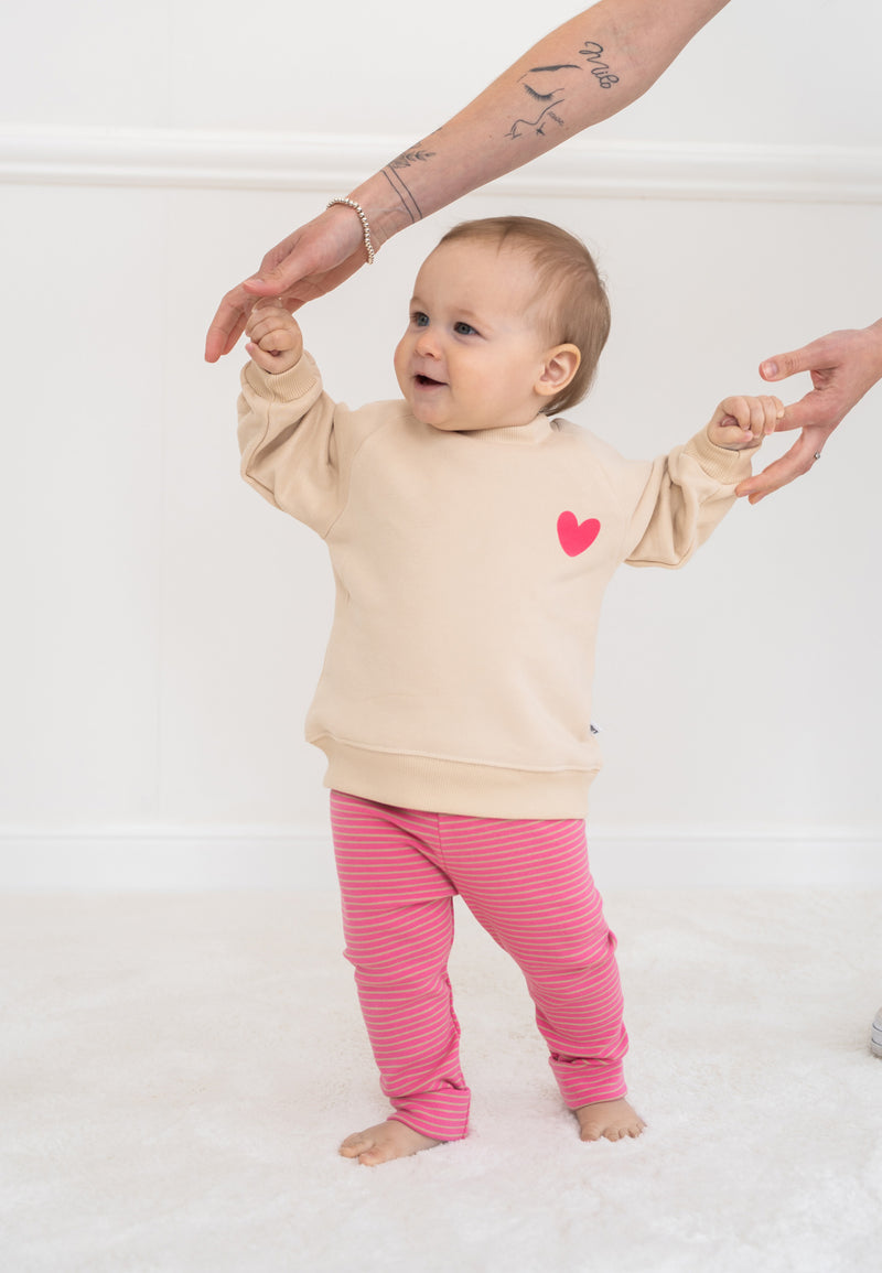 2-teiliges Baby Set mit beigem Sweatshirt mit pinkem Herz-Print und pink-rosa gestreifter Leggings, flach auf weißem Hintergrund
