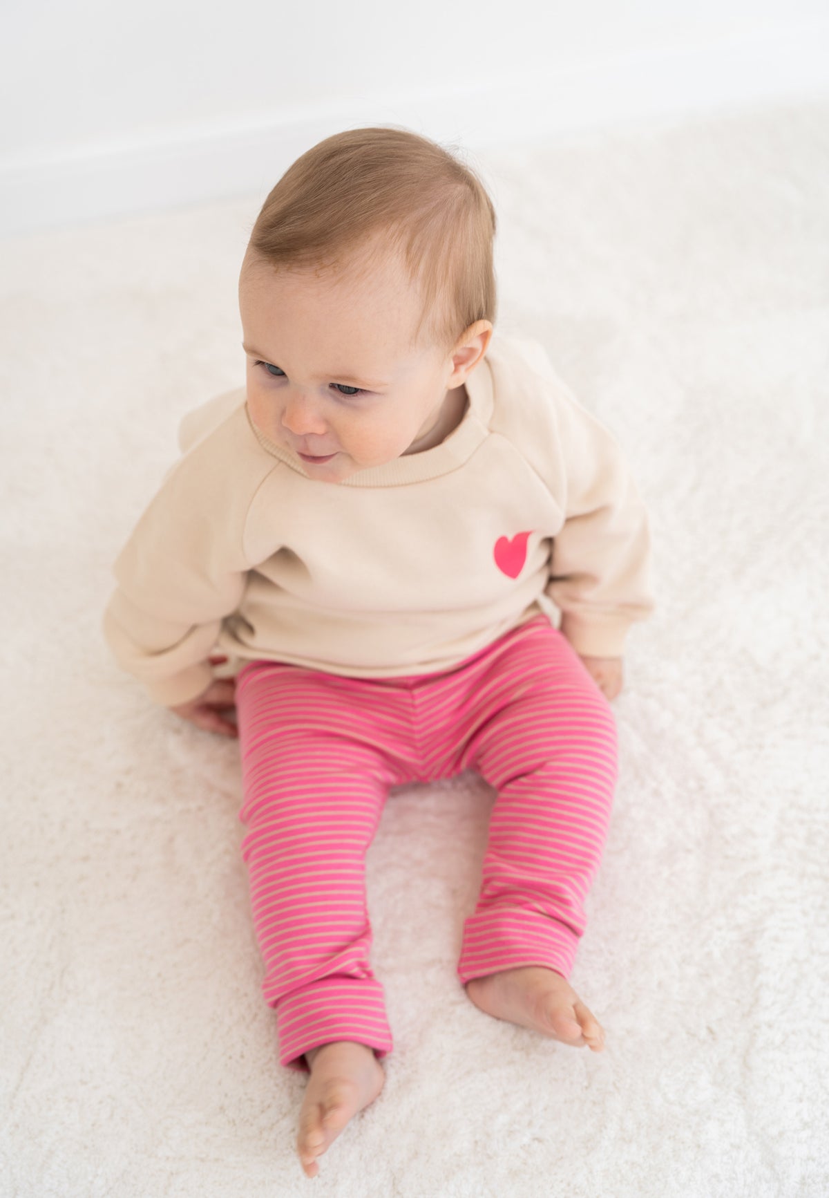 2-teiliges Baby Set mit beigem Sweatshirt mit pinkem Herz-Print und pink-rosa gestreifter Leggings, flach auf weißem Hintergrund