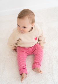 2-teiliges Baby Set mit beigem Sweatshirt mit pinkem Herz-Print und pink-rosa gestreifter Leggings, flach auf weißem Hintergrund
